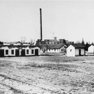 Photo en noir et blanc d'un champ avec des baraquements à un étage et une usine en arrière-plan.