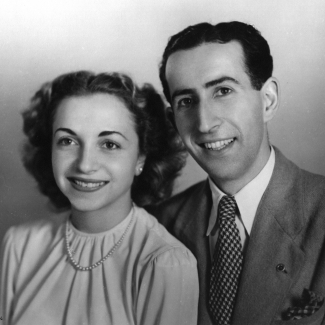 Photo en noir et blanc prise en studio d'un homme et d'une femme assis ensemble et souriant à la caméra. L'homme porte un complet et la femme une blouse avec un collier de perles. Ils ont tous deux des cheveux bruns foncés.
