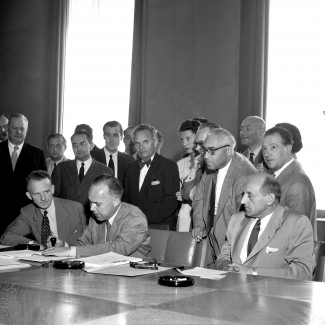 Black-and-white photograph of a group of about 25 people gathered together behind a large table in a conference room. Four men sit at the table, and one man is signing a document. The other people stand behind.