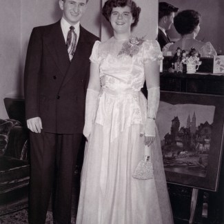 Photo en noir et blanc d'un jeune homme et d'une jeune femme se tenant debout à l'intérieur et souriant ensemble devant un mirroir. La femme porte une longue robe formelle de couleur claire ainsi qu'un petit sac à main et l'homme porte un complet.