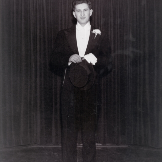 Black-and-white photograph of a young man standing in a tuxedo, in front of a dark curtain.