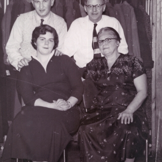 Black-and-white photograph of two men standing behind two women, who are sitting on chairs. The couple on the right appears to be older than the other couple. Both men wear shirts and ties, and the women wear dresses.