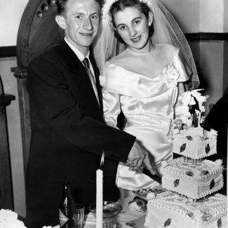 Black-and-white photograph of a man and woman standing together behind a table that holds a large, three-tiered cake. The couple, celebrating their wedding day, are cutting into the cake. They smile at the camera. The man wears a suit, and the woman wears a white silk gown with a veil.
