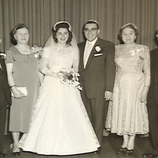 Black-and-white photograph of a group of six people, standing in a line and smiling at the camera. The couple in the middle appears to be celebrating their wedding day, with the woman dressed in a long white gown, veil, and holding a bouquet of flowers. The three men in the group wear suits, and the other two women wear dresses.