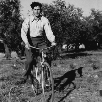 Photo en noir et blanc d'un jeune homme roulant à bicyclette à travers un champ. Il porte des pantalons foncés et une chemise à col de couleur pâle. Il y a des arbres en arrière-plan.