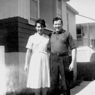 Photo en noir et blanc d'un homme et d'une femme souriant et se tenant debout dans l'allée à l'extérieur d'une maison. Il semble que ce soit l'été. La femme porte une robe à manches courtes.