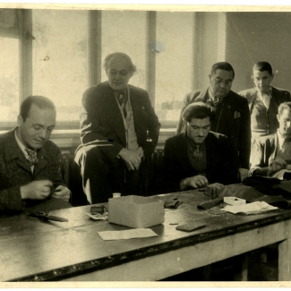 Photo en noir et blanc d'un groupe de six hommes, trois se tenant debout et trois étant asss à une table. Les trois hommes debout regardent les trois qui sont assis pendant qu'ils cousent à une table avec du fil et une aiguille.