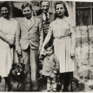 Black-and-white photograph of a man and woman standing outside the front of a house with two adolescent children, a young toddler child, and a dog.