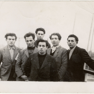 Photo en noir et blanc d'un groupe de six jeunes hommes habillés de manteaux debout ensemble sur le pont d'un navire.
