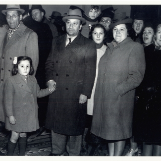 Photo en noir et blanc d'un groupe d'une douzaine de personnes rassemblés ensemble et regardant vers la caméra. Ils portent des manteaux et des chapeaux. L'homme dans la première rangée tient la main d'une jeune fille avec une boucle dans les cheveux.