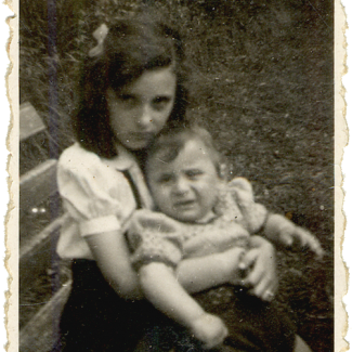 Black-and-white photograph of a girl sitting on a bench, holding a young toddler on her lap. The girl has a bow in her hair. The two siblings look at the camera, neither of them smiling.