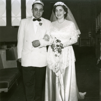 Black-and-white photograph of a man and woman, standing together arm-in-arm indoors. The couple appears to be celebrating their wedding day. The man wears a suit with a light-coloured jacket and bowtie, and the woman wears a long white gown with silk skirt, a veil, and holds a bouquet of flowers.