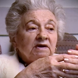 Screenshot of Holocaust survivor Mania Kay video testimony. She is sitting in front of a black background, and looking to the right of the camera. The camera shows her face and shoulders.