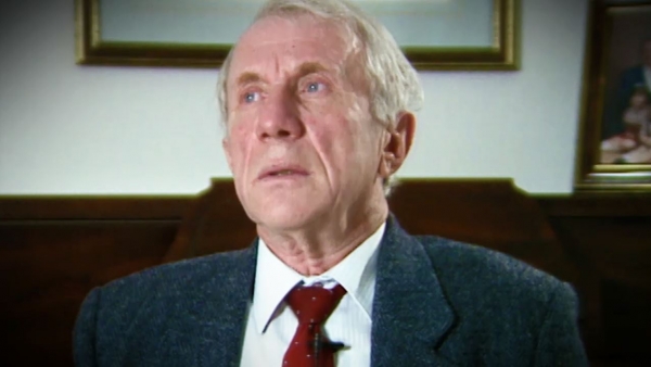 Capture d'écran du témoignage vidéo du survivant de l’Holocauste Elliott Zuckier, assis dans son salon, et regardant à la gauche de la caméra. Son visage et ses épaules sont visibles à la caméra.