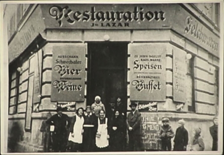 Black-and-white grainy photograph of a group of about 8 people standing in front of the entrance to a restaurant. The building has large signs on either side of the door, written in German. A couple young children look at the group from the side.