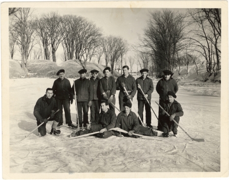 Black-and-white photograph of a hockey team of 11 men, posing together with hockey sticks on an ice rink outdoors. Trees are visible in the background.