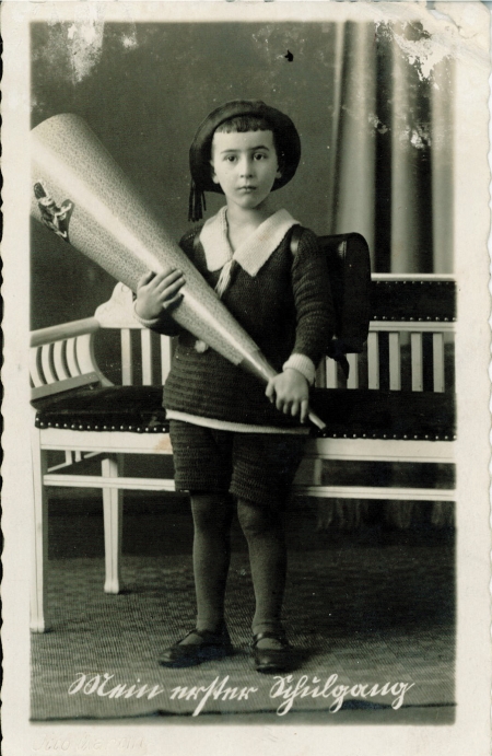 Black-and-white studio portrait photograph of a young boy wearing a school uniform and hat, looking at the camera in a pensive pose. He holds a large wooden cone-shaped object. There is white writing in German at the bottom of the image, and the edges of the photograph are slightly worn.