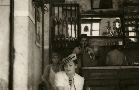 Photo en noir et blanc d'une femme assise à un café, portant une blouse blanche et un béret. La femme regarde de côté. un jeune garçon et deux hommes sont debout derrière le bar en arrière-plan.