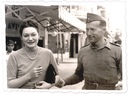 Black-and-white photograph of a woman and man outdoors. The woman holds a clutch purse under her arm and faces the camera, smiling. The man looks at the woman and holds her elbow.