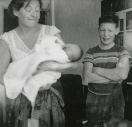 Black-and-white photograph of a young boy standing with his arms crossed and smiling beside a woman who holds a crying baby wrapped in a blanket.