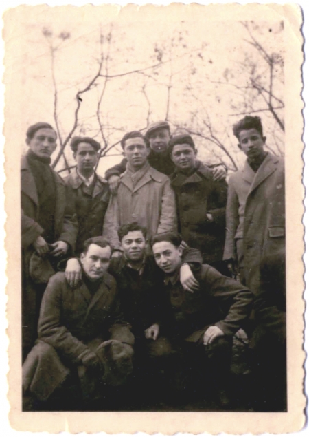 Black-and-white photograph of a group of 9 young men posing together arm-in-arm outdoors. They wear coats and there are bare trees above them in the background.