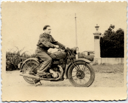 Photo en noir et blanc d'un jeune homme sur une motocyclette, regardant vers la droite de la caméra. Il y a une haie derrière lui et un bâtiment en arrière-plan.