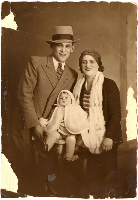 Photo de couleur sépia d'un homme et d'une femme prenant la pose avec un bébé pour un portrait. L'homme porte un chapeau ainsi que costume et cravate, et la femme porte aussi un chapeau. Le bébé porte une cape blanche. Les bords de la photo sont un peu usés et déchirés.