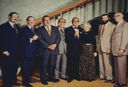 Colour photograph of a group of eight people standing together indoors in a line, smiling at the camera. The seven men wear suits, and the woman in the group wears a long dress.