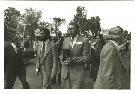 Black-and-white photograph of four men leading a march outdoors. The man in the centre is carrying a rectangular box. A group of people follows them in the background.
