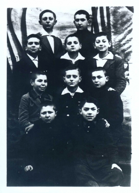Photo en noir et blanc d'un groupe de dix jeunes garçons, répartis en quatre rangs de deux ou trois. Les garçons regardent la caméra, la plupart d'entre eux portent des chemises à col sous leur veston.