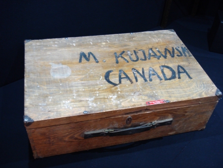 Colour photograph of a rectangular wooden suitcase with the words ‘M KUJAWSK, CANADA’ painted on the top of the case.