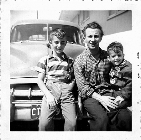 Black-and-white photograph of a man and his two boys, sitting on the front of a vintage car outdoors. The man and his sons smile at the camera.