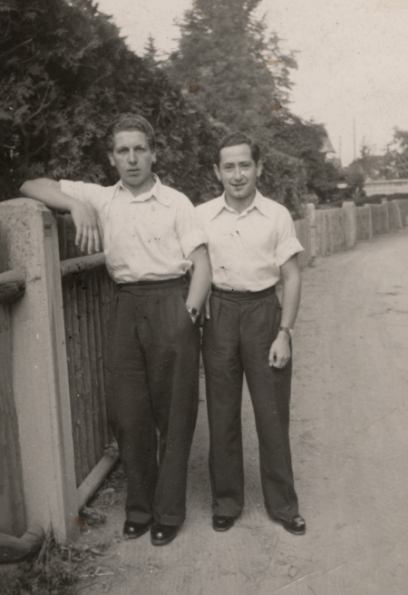 Black-and-white photograph of two young men standing together on a road, one of them leaning against a fence. They wear white collar, short sleeved shirts, and dark trousers.