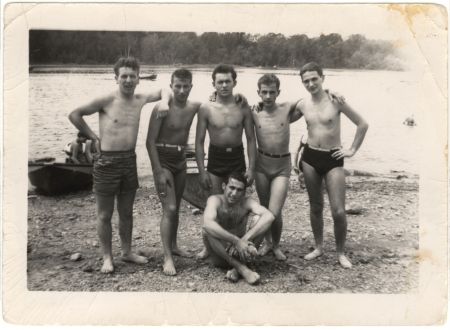 Black-and-white photograph of a group of 6 teenage boys standing arm-in-arm on a beach, with water in the background. The young men wear bathing suits, and one of them sits on the ground.