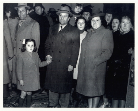 Black-and-white photograph of a group of about a dozen people standing together and looking at the camera. They wear coats and hats. The man in the front row holds the hand of a young girl with a bow in her hair.