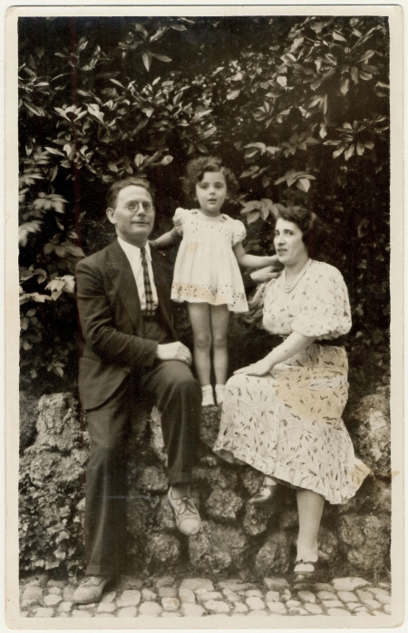 Black-and-white photograph of a man and woman sitting outdoors on a rock wall, with a young girl standing between on the wall between them, leaning on their shoulders for support. The woman and her daughter wear dresses, and the man is in a suit.