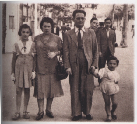 Sepia-toned photograph of a man and woman, holding the hands of their two children, standing on either side of them. The family of four is outside on a street or walkway, with people walking in the background.