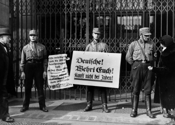 Photo en noir et blanc de trois hommes en uniforme militaire, se tenant devant une devanture de commerce fermée avec des portes en fer. Deux hommes tiennent des pancartes rédigées en allemand. L'autre soldat discute avec un civil.
