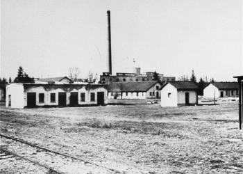 Black-and-white photograph of a field with one-storey barracks and a factory in the far background.