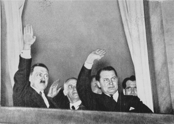 Black-and-white photograph of two men in waving out a window. The photograph is taken from below, outside. The man on the left has a moustache, and there are men behind them in the background.
