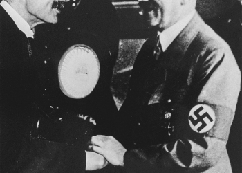 Black-and-white photograph of two men smiling and shaking hands. Both men have moustaches, and one man wears a lighter suit with a swastika badge on his arm.