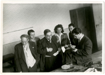 Black-and-white photograph of a group of six people, five men and one woman, standing in a room. One man is smoking a pipe and observing another man sitting on a table, hand sewing fabric.