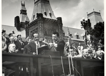 Black-and-white photograph of a man in a suit speaking into two microphones at a podium outdoors. He is surrounded by a group of people, some sitting, with a large building in the background.