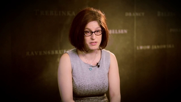 Screenshot from Adara Goldberg interview. She is sitting in front of a grey wall with various camps names written on it. Her face and shoulders are visible at the camera.