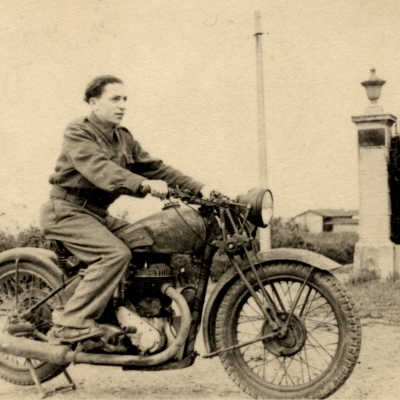 Black-and-white photograph of a young man posing on the back of a motorcycle, looking forwards towards the right of the camera. There is a hedge behind him and a building in the distant background.