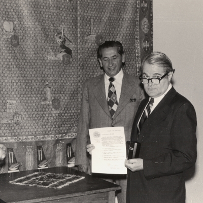 Photo en noir et blanc avec une bordure blanche sur laquelle il y a quatre hommes portant des complets rassemblés debout autour d'une table à l'intérieur. L'homme à droite tient un document et une petite urne.
