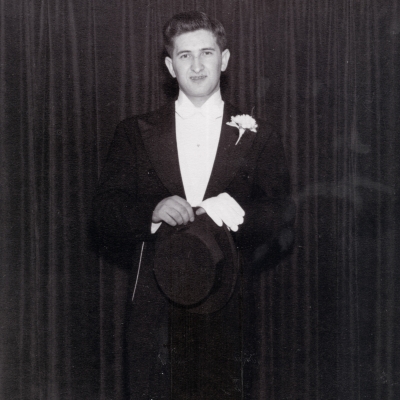 Black-and-white photograph of a young man standing in a tuxedo, in front of a dark curtain.