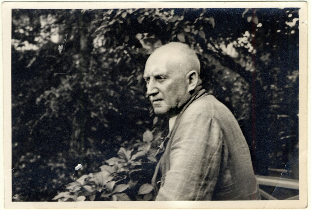 Photo en noir et blanc d'un homme âgé posant de profil à l'extérieur, entouré de feuilles et de feuillage. Il regarde au loin.