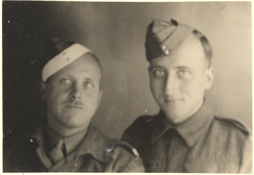 Black-and-white photograph of two men dressed in military uniform, smiling at the camera. The man on the left has a moustache.