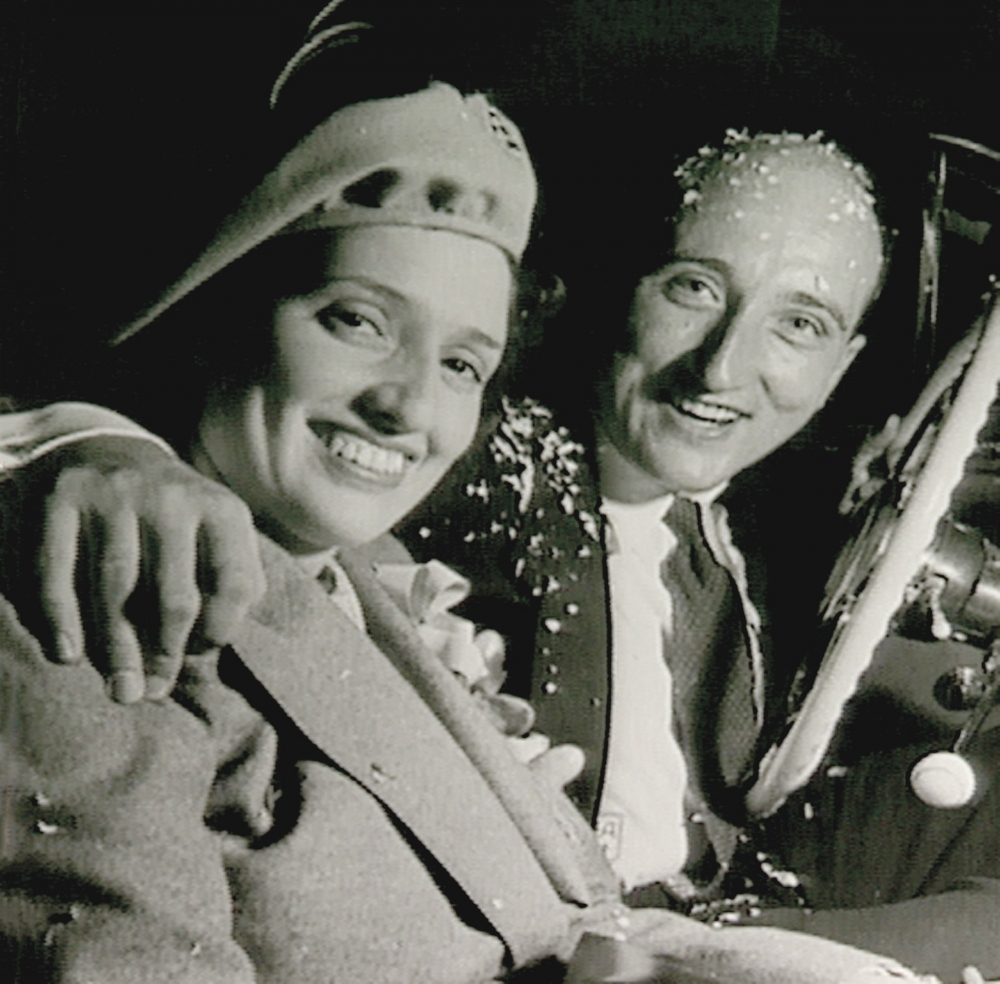 Black-and-white square-shaped photograph of a man and woman sitting together in a car, smiling. The man is covered in confetti and has his arm around the woman.